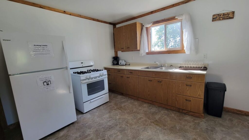 A bright kitchen area, with a fridge, gas stove, double sink, and bright window. Plenty of storage in the wooden-toned cupboards and drawers.