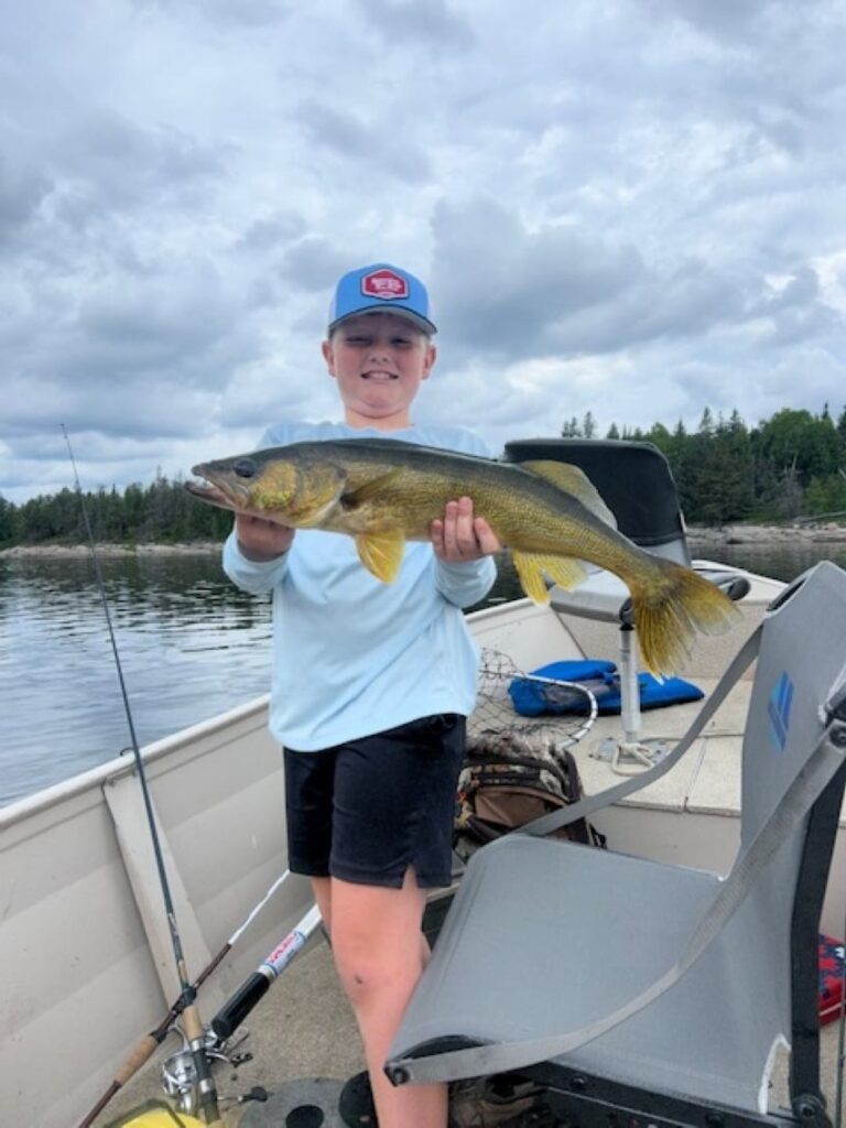 Young fisherman wearing a Separation Lake Lodge hat while holding a fish in a boat with cloudy skies on a lake and in the background you can see rocky shores with trees.