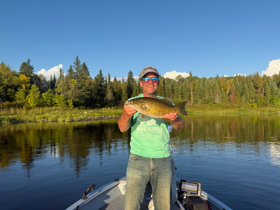 Separation Lake Lodge fisherman holding up his smallmouth bass for a picture on a sunny day while he sits in the captain seat of the boat with the lake and forest behind him